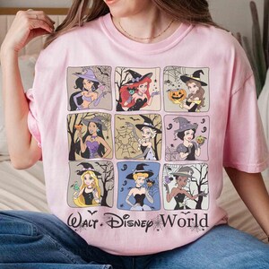May include: A pale pink t-shirt with nine square illustrations of Disney princesses in witch costumes. The shirt features the text "Walt Disney World" in black. The princesses are depicted in various poses, with Halloween themes.