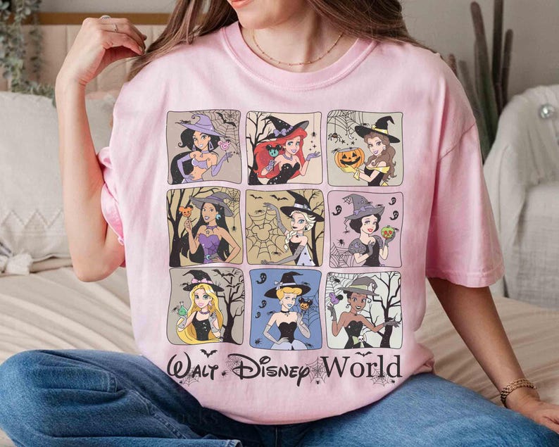 May include: A pale pink t-shirt with nine square illustrations of Disney princesses in witch costumes. The shirt features the text "Walt Disney World" in black. The princesses are depicted in various poses, with Halloween themes.