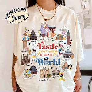 May include: Ivory-colored t-shirt with a colorful graphic featuring Disney characters and landmarks from around the world. The text "Tastle your way around the World" is displayed in a playful font. The shirt is labeled "Comfort Colors".