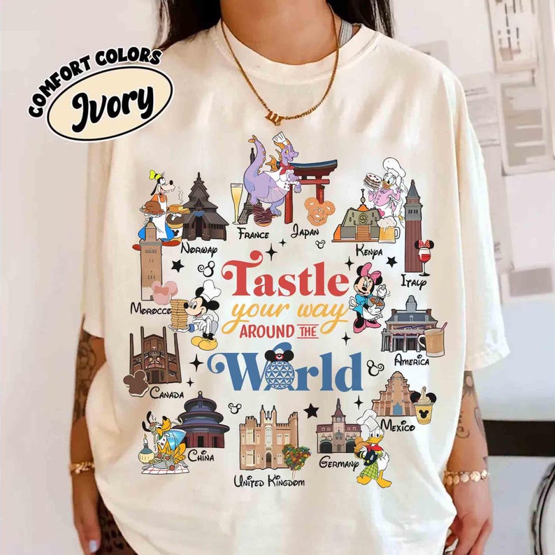 May include: Ivory-colored t-shirt with a colorful graphic featuring Disney characters and landmarks from around the world. The text "Tastle your way around the World" is displayed in a playful font. The shirt is labeled "Comfort Colors".