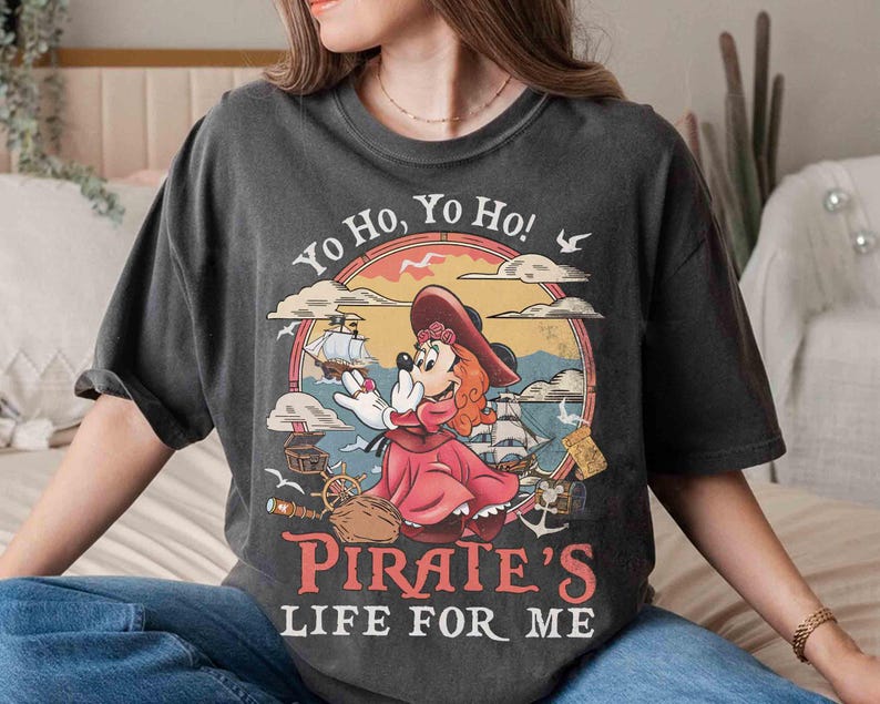 May include: Dark gray t-shirt featuring a cartoon pirate mouse character in a red dress and hat, with the text "Yo Ho, Yo Ho! Pirate's Life For Me." The design includes a ship, treasure chest, and seagulls, set within a circular frame.