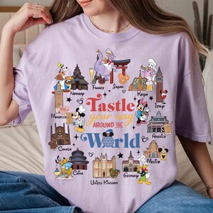 May include: A light purple t-shirt featuring a colorful graphic with the text "Tastle your way around the World". The graphic includes cartoon characters and landmarks from various countries, such as France, Japan, and Italy.