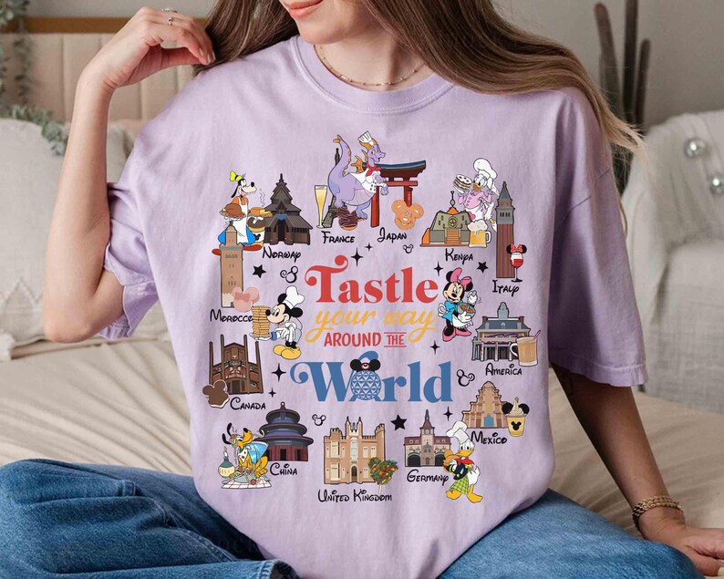 May include: A light purple t-shirt featuring a colorful graphic with the text "Tastle your way around the World". The graphic includes cartoon characters and landmarks from various countries, such as France, Japan, and Italy.