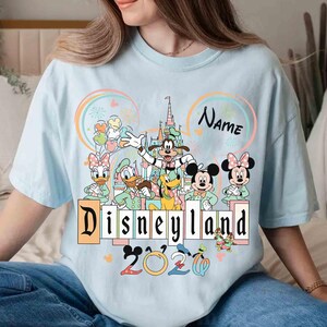 May include: Light blue t-shirt featuring a Disneyland graphic with Mickey Mouse, Minnie Mouse, Goofy, Donald Duck, and Daisy Duck. The design includes the text "Disneyland 2020" and a space for a name. The shirt has a crew neck.