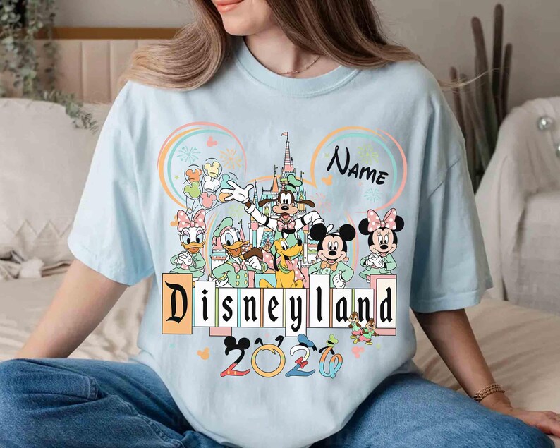 May include: Light blue t-shirt featuring a Disneyland graphic with Mickey Mouse, Minnie Mouse, Goofy, Donald Duck, and Daisy Duck. The design includes the text "Disneyland 2020" and a space for a name. The shirt has a crew neck.