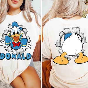 May include: Cream-colored t-shirt featuring a cartoon image of Donald Duck bursting through a torn hole, with the name "DONALD" below. The back of the shirt shows Donald Duck's rear and feet. The design includes blue, white, red, and yellow.