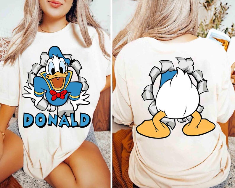 May include: Cream-colored t-shirt featuring a cartoon image of Donald Duck bursting through a torn hole, with the name "DONALD" below. The back of the shirt shows Donald Duck's rear and feet. The design includes blue, white, red, and yellow.