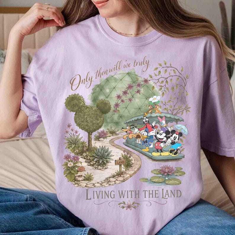 May include: A light purple t-shirt featuring a whimsical illustration with the text "Only then will we truly" and "Living with the Land." The design includes cartoon characters, topiaries, and a scenic landscape.