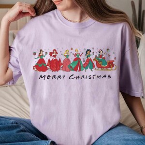 May include: A light purple t-shirt featuring a graphic of several princesses in festive holiday attire. The princesses are in red, green, and pink dresses. The text "MERRY CHRISTMAS" is printed below the graphic.
