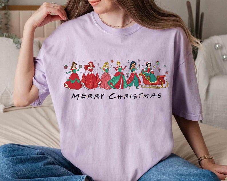 May include: A light purple t-shirt featuring a graphic of several princesses in festive holiday attire. The princesses are in red, green, and pink dresses. The text "MERRY CHRISTMAS" is printed below the graphic.