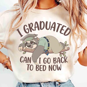 May include: Cream-colored t-shirt with the text "I GRADUATED CAN I GO BACK TO BED NOW." The graphic features a cartoon sloth wearing a graduation cap and gown, lying on a branch. The sloth is light brown and gray.