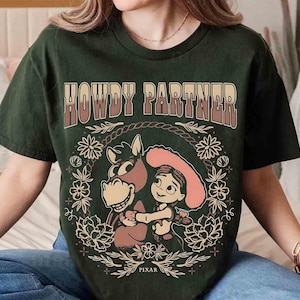Puede incluir: Una camiseta de manga corta de color verde oscuro con el texto HOWDY PARTNER en una fuente degradada sobre un gr&aacute;fico de una vaquera de dibujos animados y un caballo. El dise&ntilde;o est&aacute; rodeado por una corona floral e incluye la palabra PIXAR.