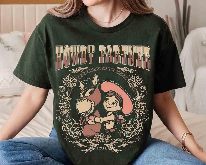Puede incluir: Una camiseta de manga corta de color verde oscuro con el texto HOWDY PARTNER en una fuente degradada sobre un gr&aacute;fico de una vaquera de dibujos animados y un caballo. El dise&ntilde;o est&aacute; rodeado por una corona floral e incluye la palabra PIXAR.