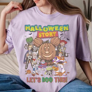 Puede incluir: Camiseta lila claro con un dise&ntilde;o de Halloween. El dise&ntilde;o presenta una calabaza sonriente, personajes de dibujos animados disfrazados de Halloween y el texto "HALLOWEEN STORY" y "LET'S BOO THIS!"