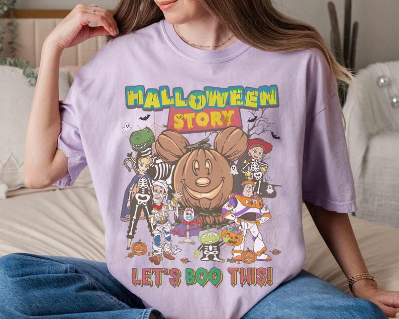 Puede incluir: Camiseta lila claro con un dise&ntilde;o de Halloween. El dise&ntilde;o presenta una calabaza sonriente, personajes de dibujos animados disfrazados de Halloween y el texto "HALLOWEEN STORY" y "LET'S BOO THIS!"
