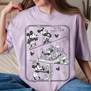 Może przedstawiać: Jasnofioletowa koszulka z czarno-białą grafiką Myszki Miki ze Steamboat Willie. Grafika przedstawia Myszkę Miki w r&oacute;żnych pozach, napis "Steamboat Willie EST. 1928" i sylwetki Myszki Miki.