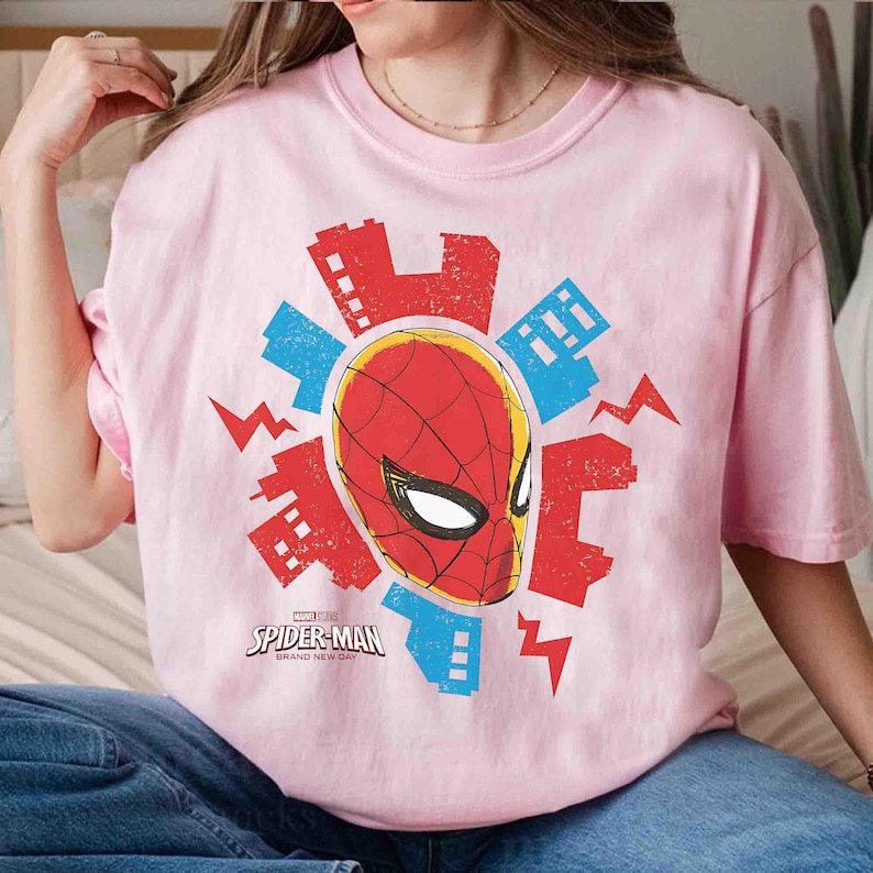 Puede incluir: Una camiseta de cuello redondo rosa claro con un gr&aacute;fico central de la m&aacute;scara de Spider-Man rodeada por siluetas de edificios rojos y azules y formas de rayos. El logotipo de Marvel Spider-Man Brand New Day est&aacute; impreso debajo.