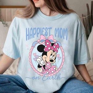 Pode incluir: Camiseta azul claro com Minnie Mouse em vestido e la&ccedil;o de bolinhas rosa. O texto "Happiest Mom on Earth" est&aacute; em azul. O desenho &eacute; emoldurado em um oval rosa e azul.