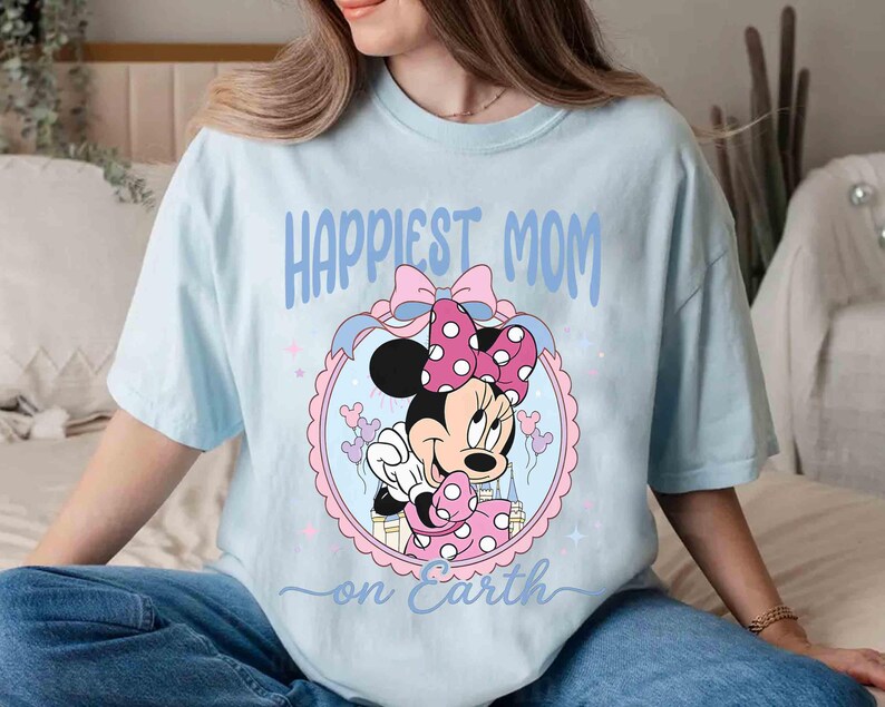 Pode incluir: Camiseta azul claro com Minnie Mouse em vestido e la&ccedil;o de bolinhas rosa. O texto "Happiest Mom on Earth" est&aacute; em azul. O desenho &eacute; emoldurado em um oval rosa e azul.