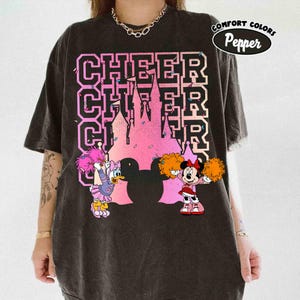 May include: Black t-shirt with a pink and purple castle graphic and the word "CHEER" in a collegiate font. Daisy Duck and Minnie Mouse are depicted as cheerleaders with pom-poms. The shirt has a "Comfort Colors" and "Pepper" logo.