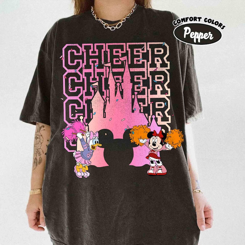 May include: Black t-shirt with a pink and purple castle graphic and the word "CHEER" in a collegiate font. Daisy Duck and Minnie Mouse are depicted as cheerleaders with pom-poms. The shirt has a "Comfort Colors" and "Pepper" logo.