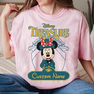 K&ouml;nnte beinhalten: Hellrosa T-Shirt mit einer Minnie Mouse Grafik in Kapit&auml;nsuniform, dem Text "Disney Treasure" und Platz f&uuml;r einen individuellen Namen. Das Design beinhaltet eine Schiffskontur.