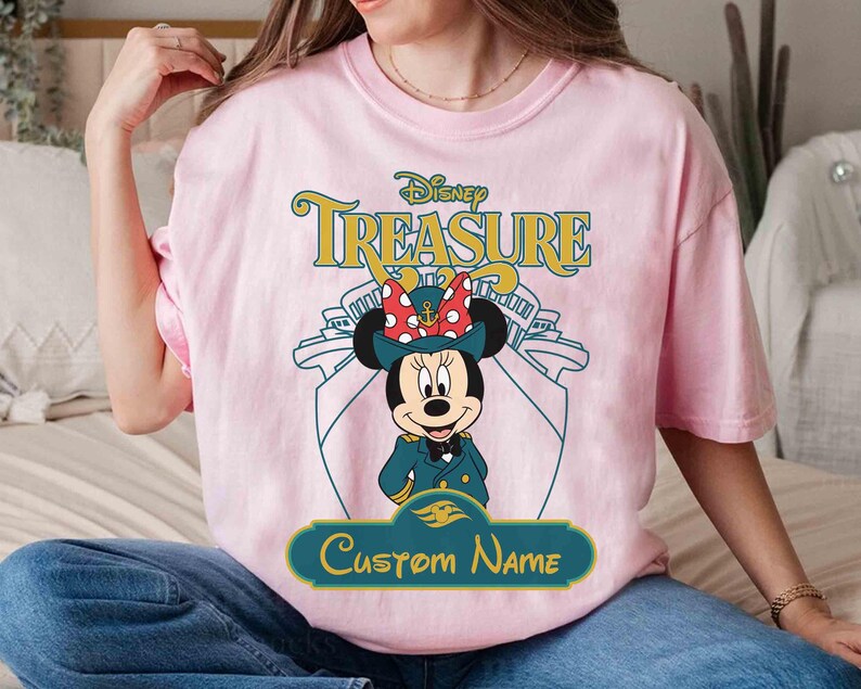 K&ouml;nnte beinhalten: Hellrosa T-Shirt mit einer Minnie Mouse Grafik in Kapit&auml;nsuniform, dem Text "Disney Treasure" und Platz f&uuml;r einen individuellen Namen. Das Design beinhaltet eine Schiffskontur.