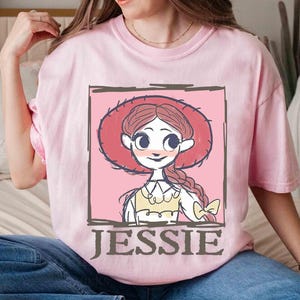 May include: A light pink t-shirt featuring a cartoon illustration of Jessie from Toy Story. The illustration is framed in a dark brown border and includes the name "JESSIE" in a matching font. The character is wearing a red hat.