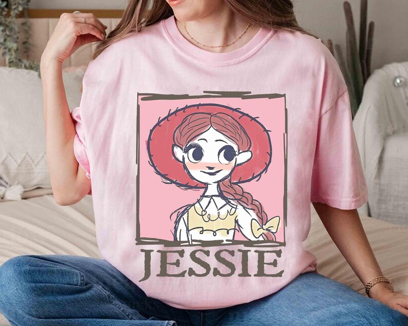 May include: A light pink t-shirt featuring a cartoon illustration of Jessie from Toy Story. The illustration is framed in a dark brown border and includes the name "JESSIE" in a matching font. The character is wearing a red hat.