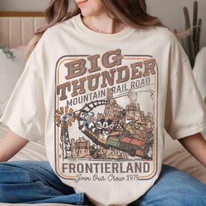 May include: Cream-colored t-shirt featuring a vintage-style graphic of the Big Thunder Mountain Railroad ride. The design includes the text "Big Thunder Mountain Railroad Frontierland Join Our Crew 1979" with cartoon characters and a train.