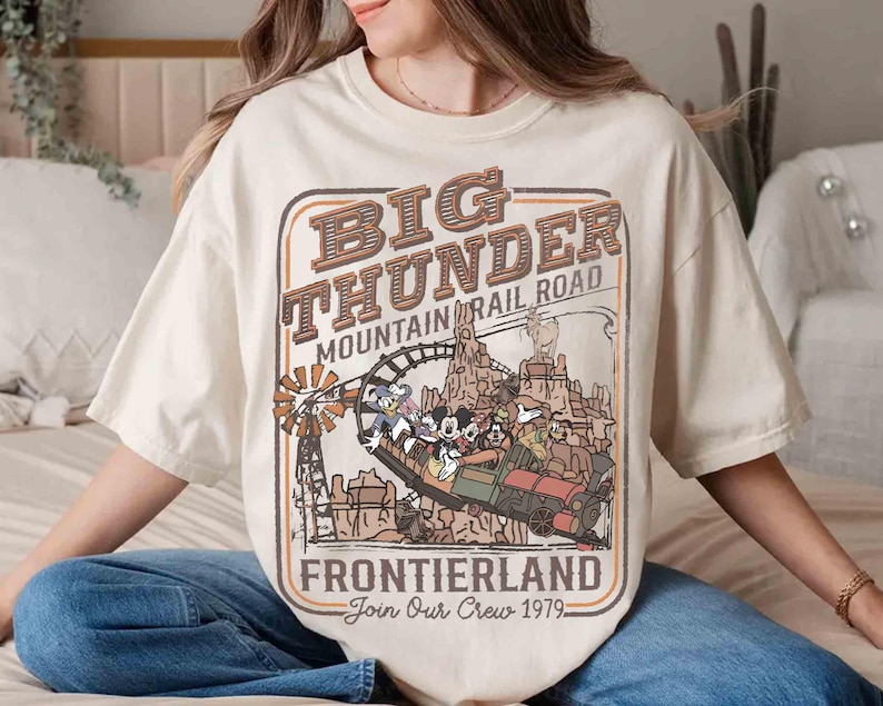 May include: Cream-colored t-shirt featuring a vintage-style graphic of the Big Thunder Mountain Railroad ride. The design includes the text "Big Thunder Mountain Railroad Frontierland Join Our Crew 1979" with cartoon characters and a train.