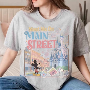 May include: A gray t-shirt featuring the text "Meet Me On Main Street" in colorful lettering. The design includes illustrations of a castle, Mickey Mouse, and other theme park elements. The shirt is a casual wear item.