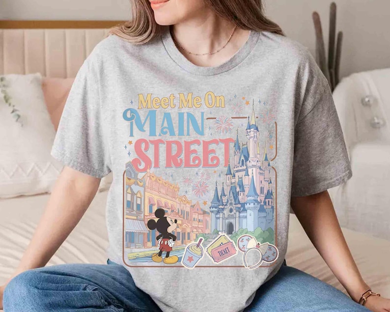 May include: A gray t-shirt featuring the text "Meet Me On Main Street" in colorful lettering. The design includes illustrations of a castle, Mickey Mouse, and other theme park elements. The shirt is a casual wear item.