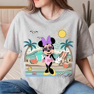 K&ouml;nnte beinhalten: Ein graues T-Shirt mit einem Cartoon-Design von Minnie Mouse am Strand. Minnie tr&auml;gt herzf&ouml;rmige Sonnenbrillen, eine lila Schleife und ein rosa Kleid. Das Design umfasst Palmen, eine Sonne und den Text "Minnie Mouse XOXO".