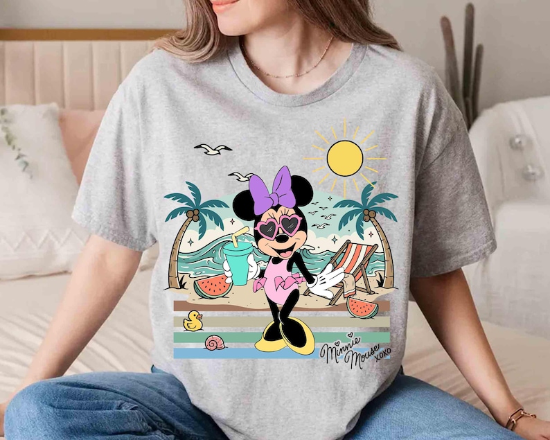 K&ouml;nnte beinhalten: Ein graues T-Shirt mit einem Cartoon-Design von Minnie Mouse am Strand. Minnie tr&auml;gt herzf&ouml;rmige Sonnenbrillen, eine lila Schleife und ein rosa Kleid. Das Design umfasst Palmen, eine Sonne und den Text "Minnie Mouse XOXO".