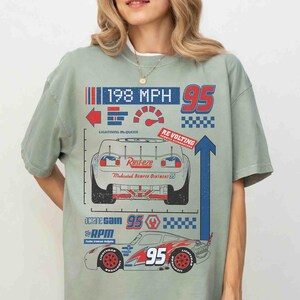 K&ouml;nnte beinhalten: Ein salbeigr&uuml;nes T-Shirt mit einem Grafikdruck eines Rennwagens. Das Design enth&auml;lt den Text 198 MPH, 95, Lightning McQueen, Rust-eze, Re-Volting, Octane Gain und RPM. Der Druck zeigt eine R&uuml;ck- und Seitenansicht des Wagens in Rot, Blau und Wei&szlig;.