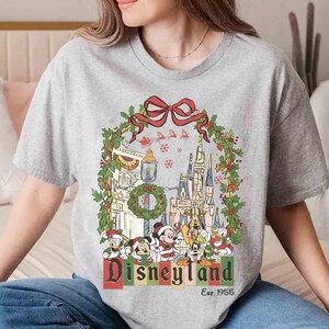 Op de afbeelding: Grijze t-shirt met een Disneyland kerstontwerp. De afbeelding toont een kasteel, Mickey Mouse, Donald Duck en andere personages, omlijst door een krans en een rode strik. De tekst "Disneyland" en "Est. 1955" zijn ook zichtbaar.