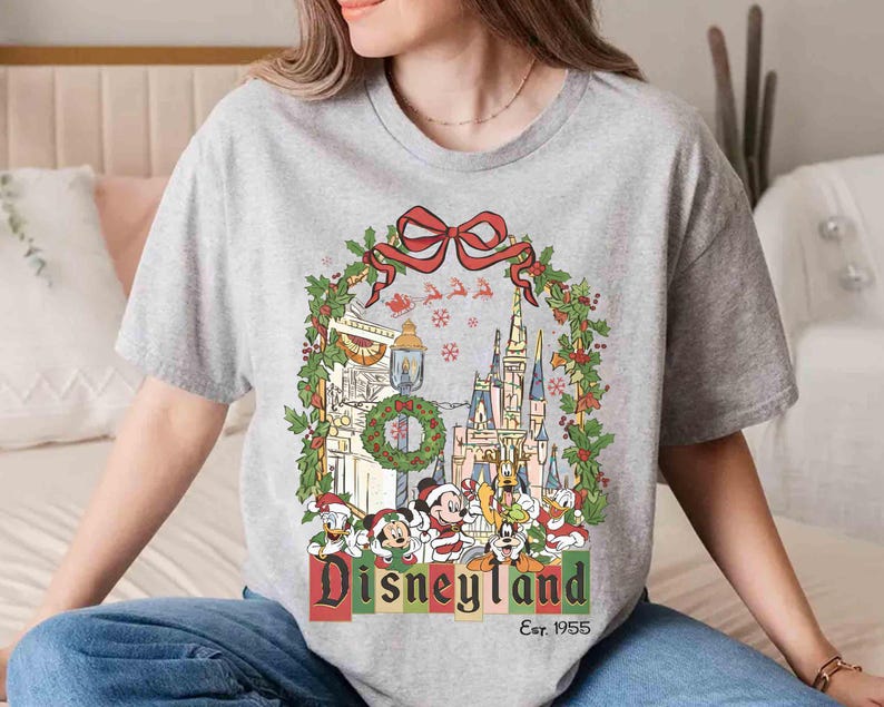 Op de afbeelding: Grijze t-shirt met een Disneyland kerstontwerp. De afbeelding toont een kasteel, Mickey Mouse, Donald Duck en andere personages, omlijst door een krans en een rode strik. De tekst "Disneyland" en "Est. 1955" zijn ook zichtbaar.