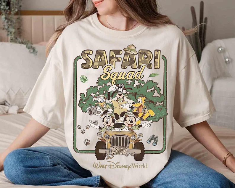 May include: A cream-coloured t-shirt with a graphic of Disney characters in a safari jeep. The design includes the text "SAFARI Squad" and "Walt Disney World". The jeep is surrounded by cartoon trees and paw prints.