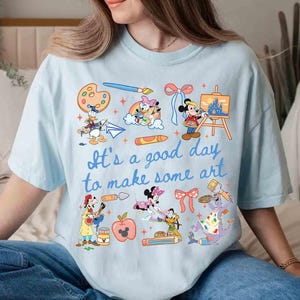 May include: Light blue t-shirt with the phrase "It's a good day to make some art" and various cartoon characters. The shirt features illustrations of paint palettes, paintbrushes, and art supplies, with characters like Mickey Mouse and Donald Duck.
