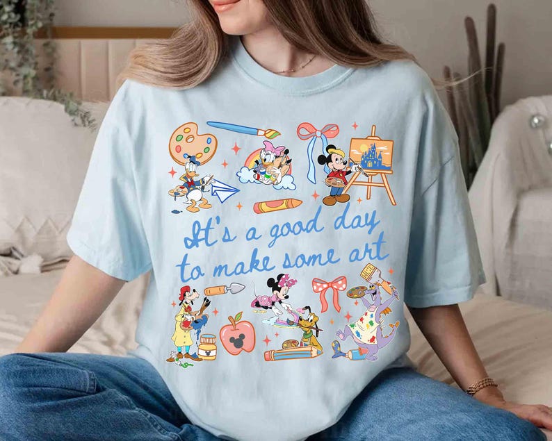 May include: Light blue t-shirt with the phrase "It's a good day to make some art" and various cartoon characters. The shirt features illustrations of paint palettes, paintbrushes, and art supplies, with characters like Mickey Mouse and Donald Duck.