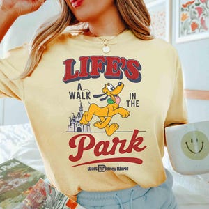May include: Yellow t-shirt with the text "LIFE'S A WALK IN THE PARK" in red and blue, featuring a cartoon Pluto character and a castle graphic. The shirt also has the "Walt Disney World" logo.