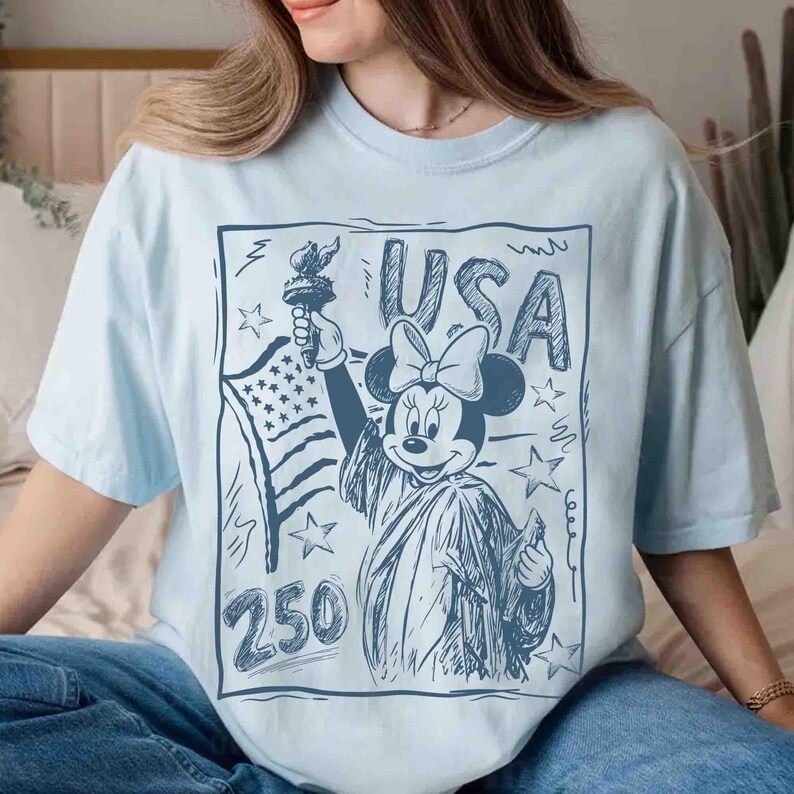 K&ouml;nnte beinhalten: Hellblaues T-Shirt mit einer Grafik von Minnie Maus als Freiheitsstatue, die eine Fackel und die amerikanische Flagge h&auml;lt. Das Wort "USA" steht &uuml;ber Minnie, mit der Zahl "250" darunter. Das Design ist in Blaut&ouml;nen gehalten.