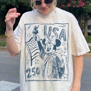 K&ouml;nnte beinhalten: Cremefarbenes T-Shirt mit einer blauen Grafik von Minnie Mouse als Freiheitsstatue, die eine Fackel und die amerikanische Flagge h&auml;lt. Das Wort "USA" und die Zahl "250" sind ebenfalls im Design enthalten.