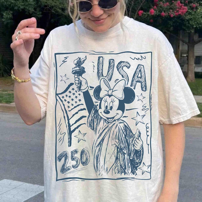 K&ouml;nnte beinhalten: Cremefarbenes T-Shirt mit einer blauen Grafik von Minnie Mouse als Freiheitsstatue, die eine Fackel und die amerikanische Flagge h&auml;lt. Das Wort "USA" und die Zahl "250" sind ebenfalls im Design enthalten.