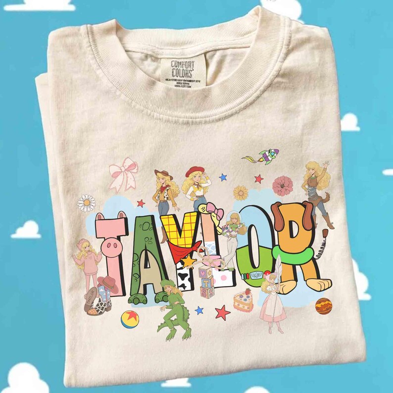 May include: A cream-colored t-shirt with a graphic print featuring the name TAYLOR in large, colorful, stylized letters. The design includes various cartoon characters, small stars, flowers, a ball, and a cake scattered around the text.
