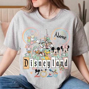 May include: A gray t-shirt featuring a Disneyland graphic with Mickey Mouse, Minnie Mouse, Donald Duck, Daisy Duck, Goofy, and Pluto. The design includes the text "Disneyland" and "2020", with a space for a name.