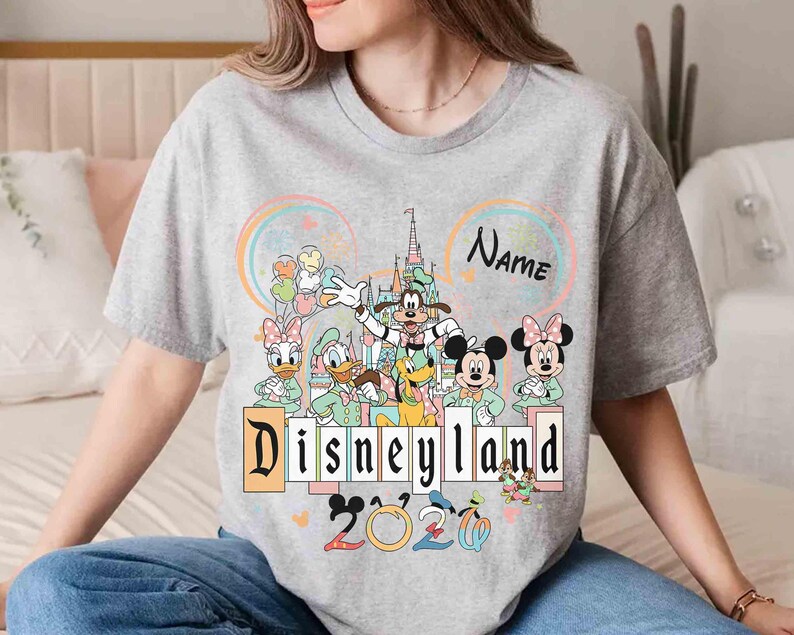 May include: A gray t-shirt featuring a Disneyland graphic with Mickey Mouse, Minnie Mouse, Donald Duck, Daisy Duck, Goofy, and Pluto. The design includes the text "Disneyland" and "2020", with a space for a name.