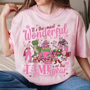 May include: A light pink t-shirt with the text "It's the most Wonderful Time of the Year" in pink script. The shirt features cartoon characters, a Christmas tree, and presents. The design is festive and holiday-themed.