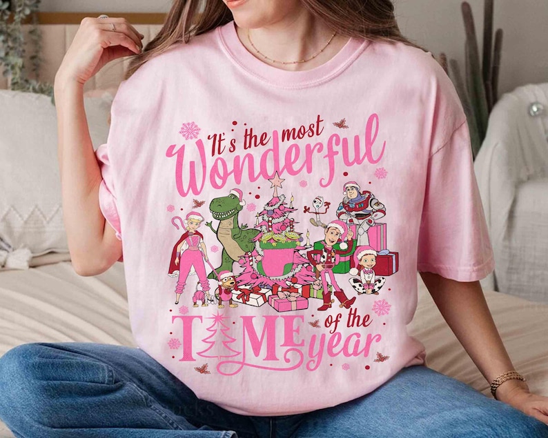 May include: A light pink t-shirt with the text "It's the most Wonderful Time of the Year" in pink script. The shirt features cartoon characters, a Christmas tree, and presents. The design is festive and holiday-themed.